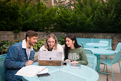 Three students sitting at an outdoor table looking at a laptop computer.
