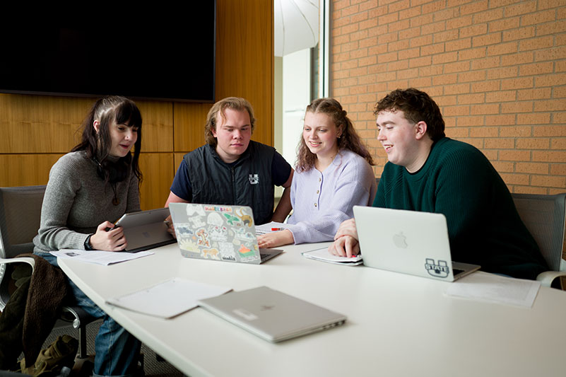 Four students working together at a table using laptops and a tablet.
