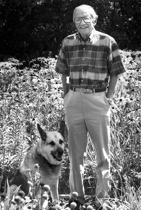 Black and white photo of Tom smiling in a sunflower field standing next to a German Shepherd