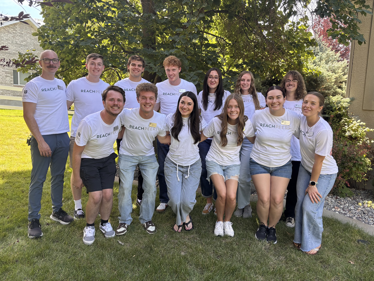 Group of  13 REACH Peer members wearing REACH Peer white t-shirts, outside.