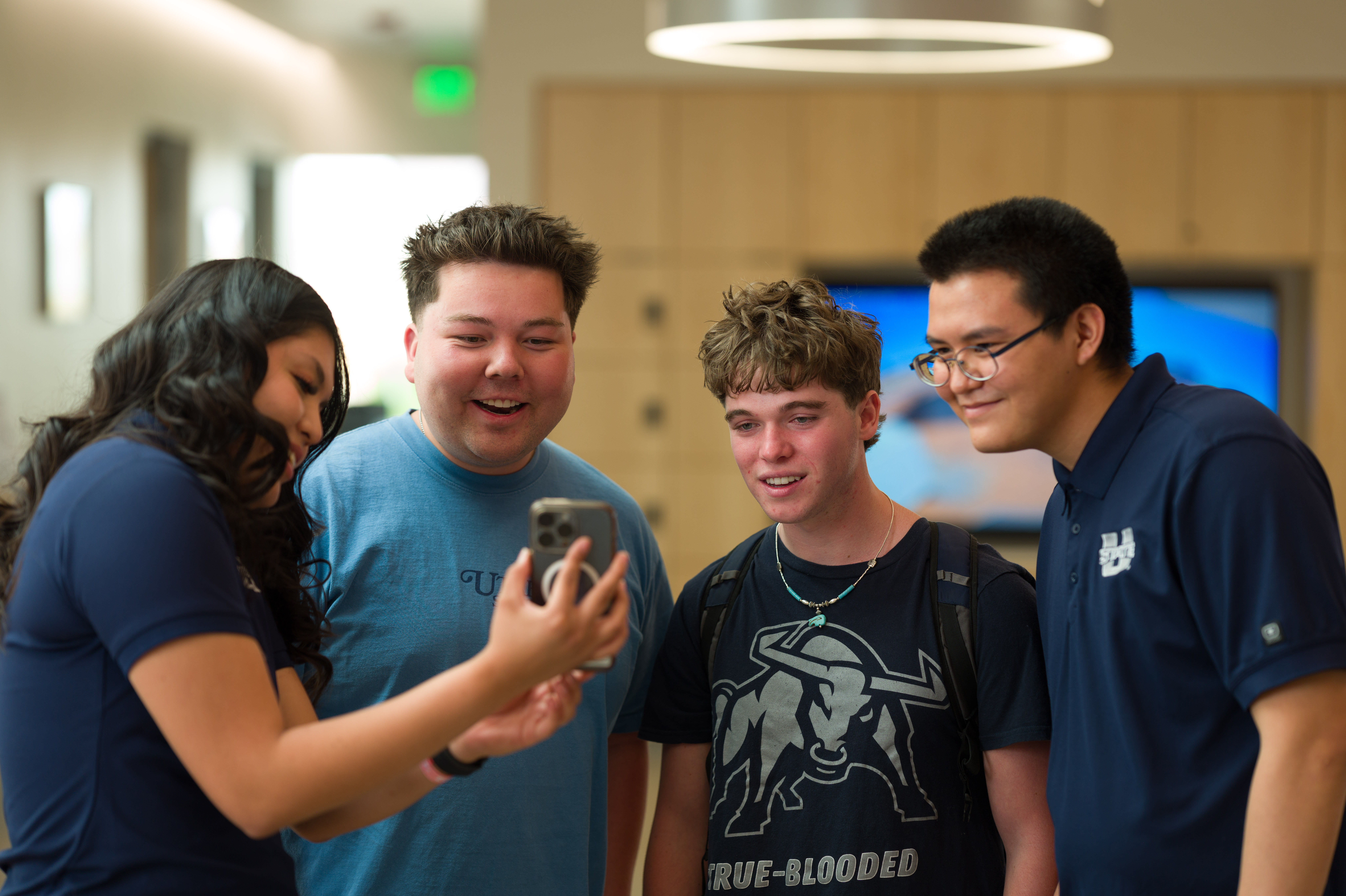 Four students standing indoors, smiling and looking at a smartphone held by one of them. They wear casual clothes, including shirts with Utah State University logos, and appear engaged in a friendly conversation.
