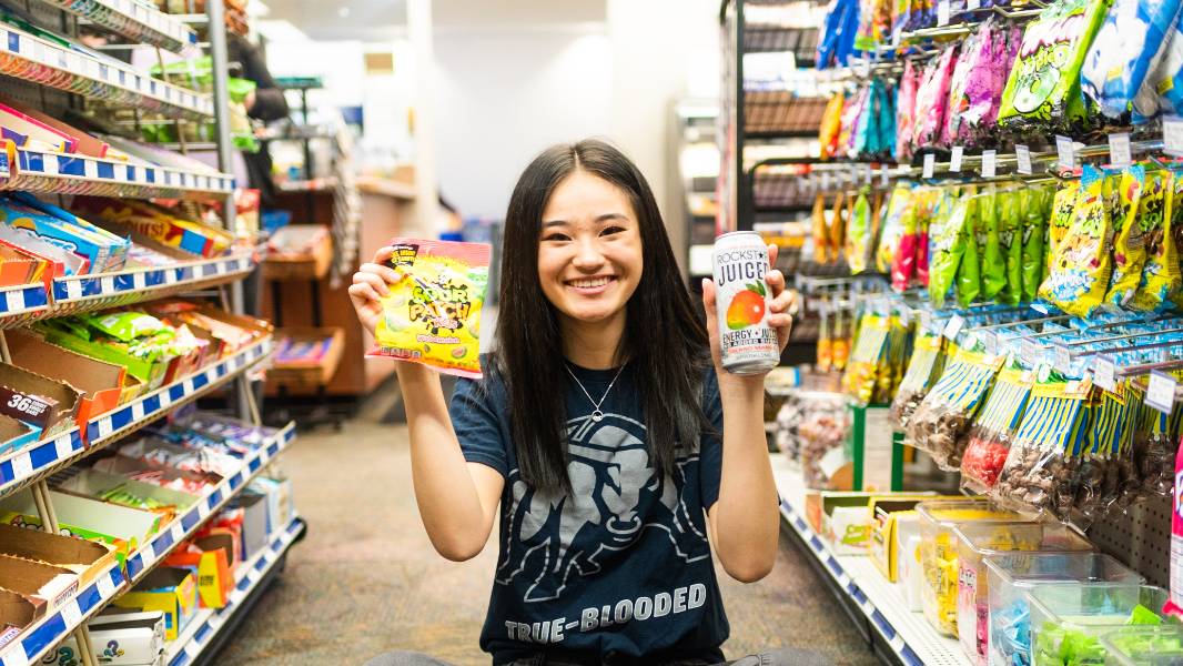 girl in an aisle full of candy. She is smiling as she holds a bag of sour patch watermelons in one hand and a rock star energy drink in the other