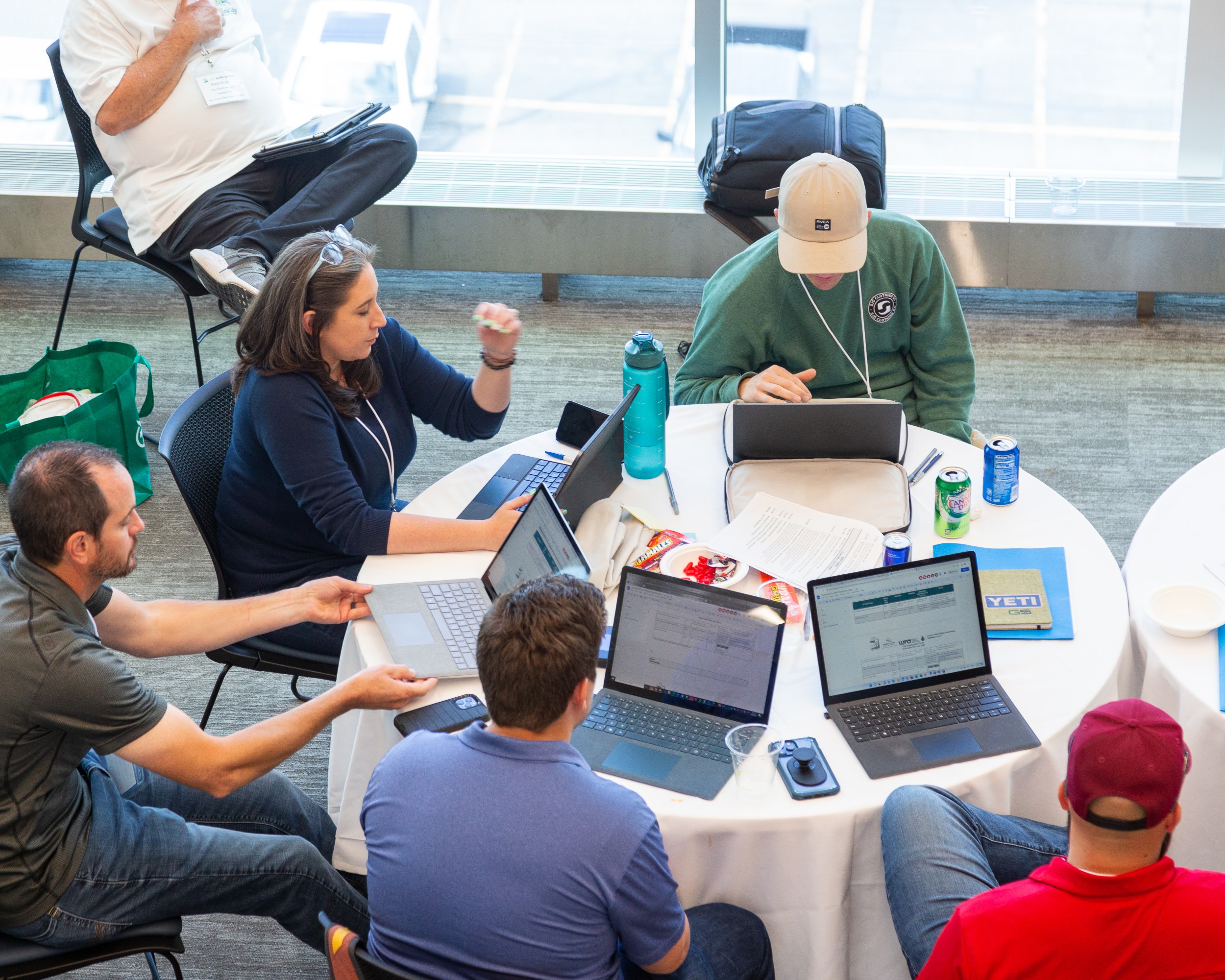 Individuals at a round table conversate and work on laptops.