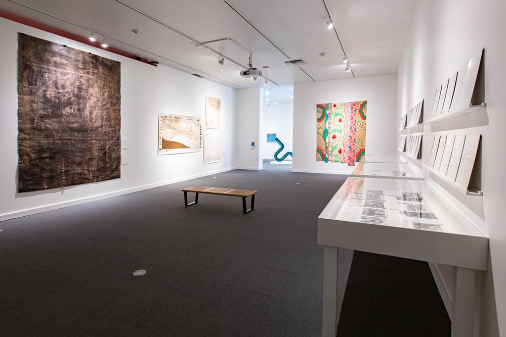 A little bench in the middle of a room with a large tapestry on the left wall and shadow box tables on the right