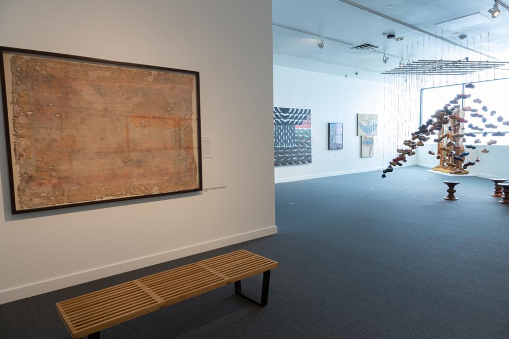A bench sits in front of a framed art piece in the distance clogs hang from the ceiling