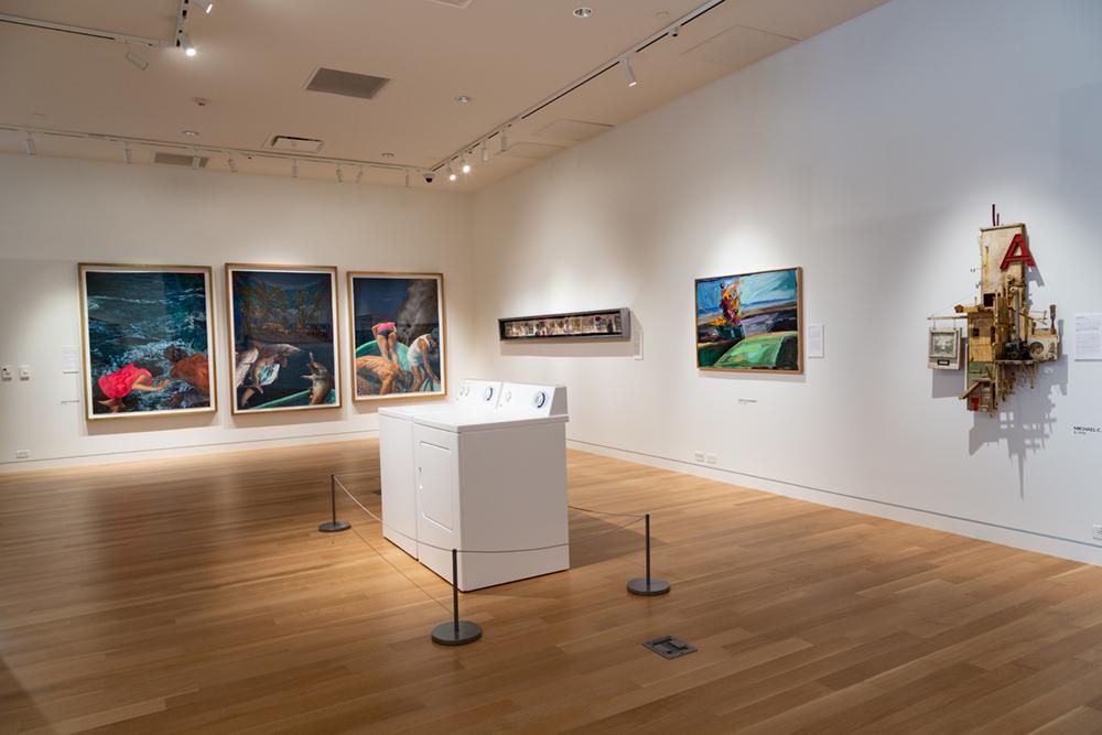 A washing machine and dryer display in the middle of the room with various art pieces hung on the wall behind