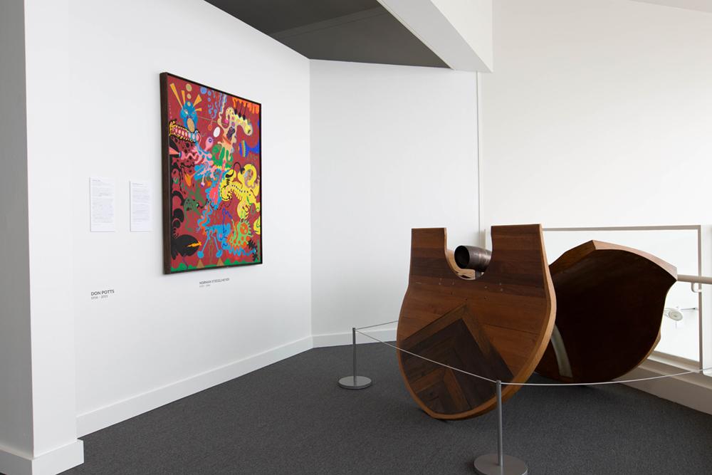 A large sculpture on the floor with ropes around it and one colorful art piece hanging behind it