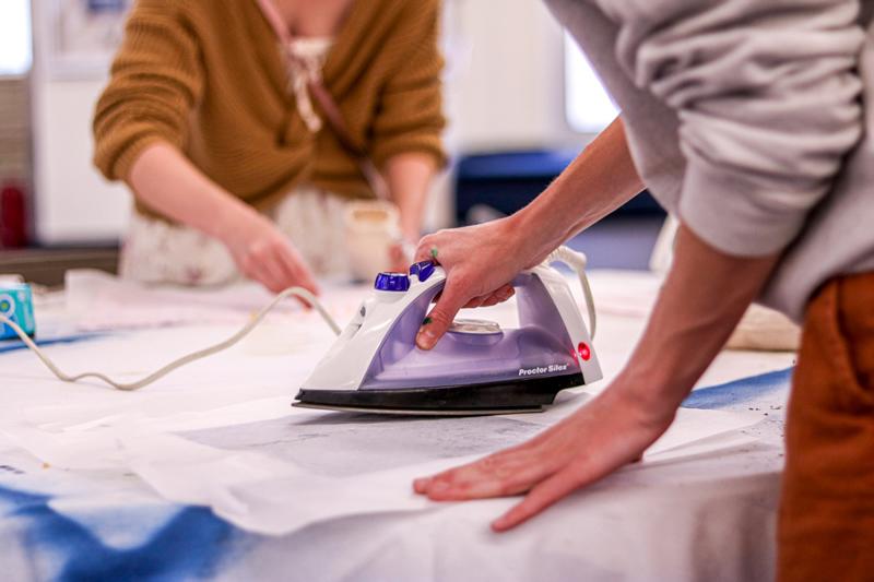 Foreground student ironing and background student sprinkling wax onto fabric.
