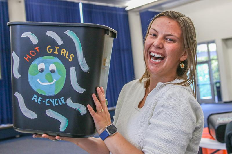 Another picture of student holding up their completed recycling bin painted with a smiling Earth surrounded by the words "Hot girls re-cycle".