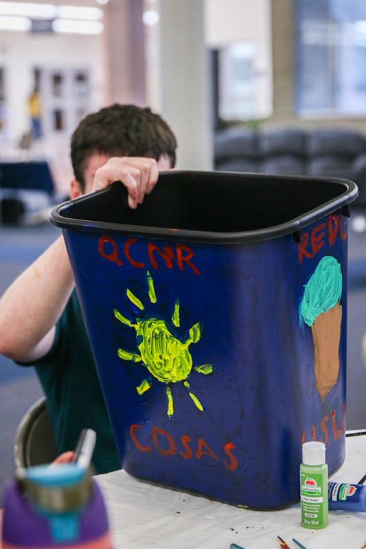 Student painting their recycling bin with a sun and the words "QCNR, COSAS, and USU".