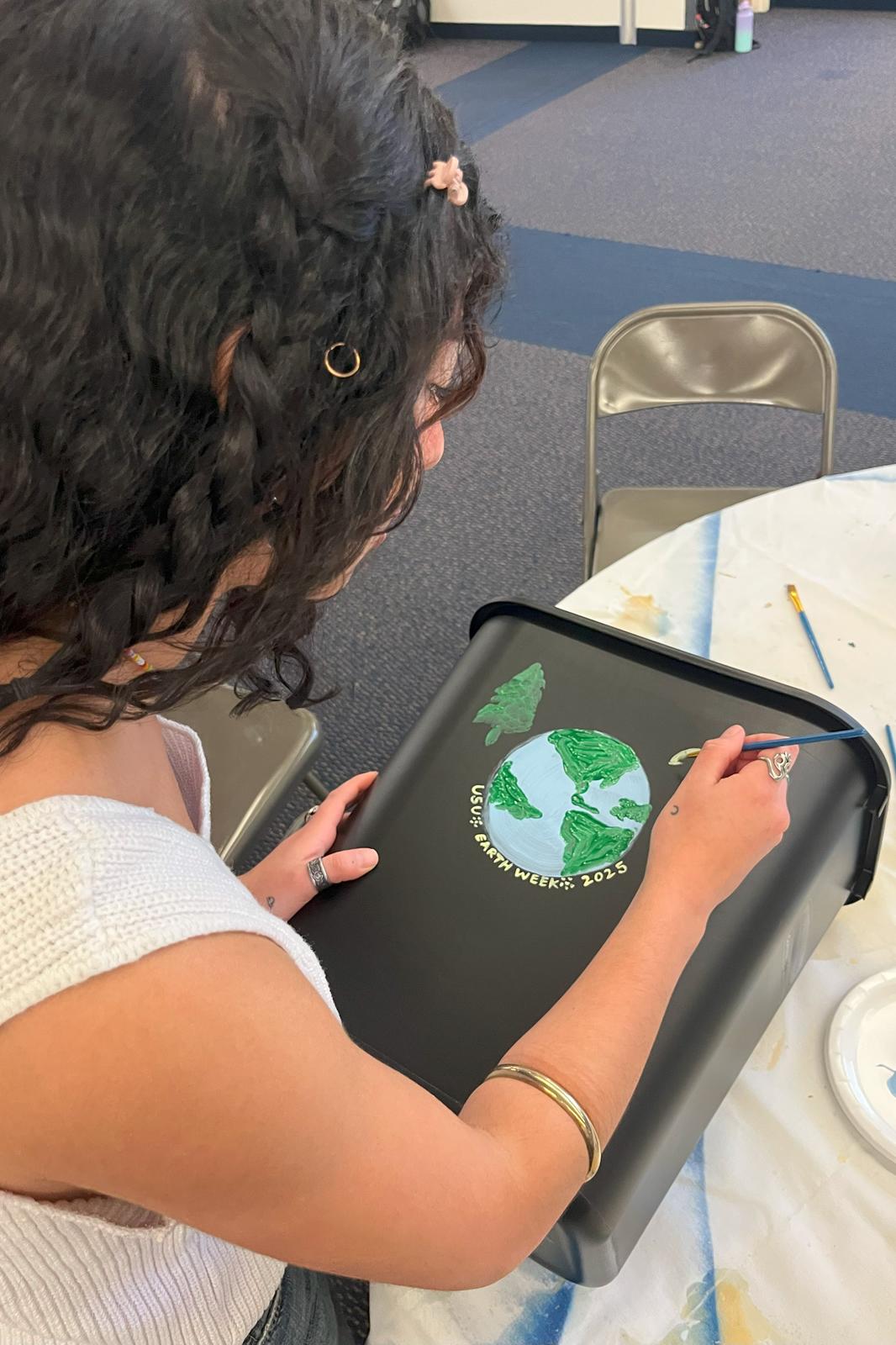 Student painting a recycling bin with a pine tree, Earth, and the words "USU Earth Week 2025".
