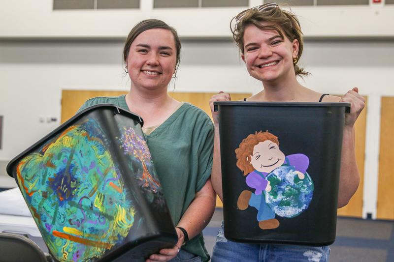 Two students holding up their completed recycling bins, one bin with colorful abstract design, the other with a person smiling and holding Earth.