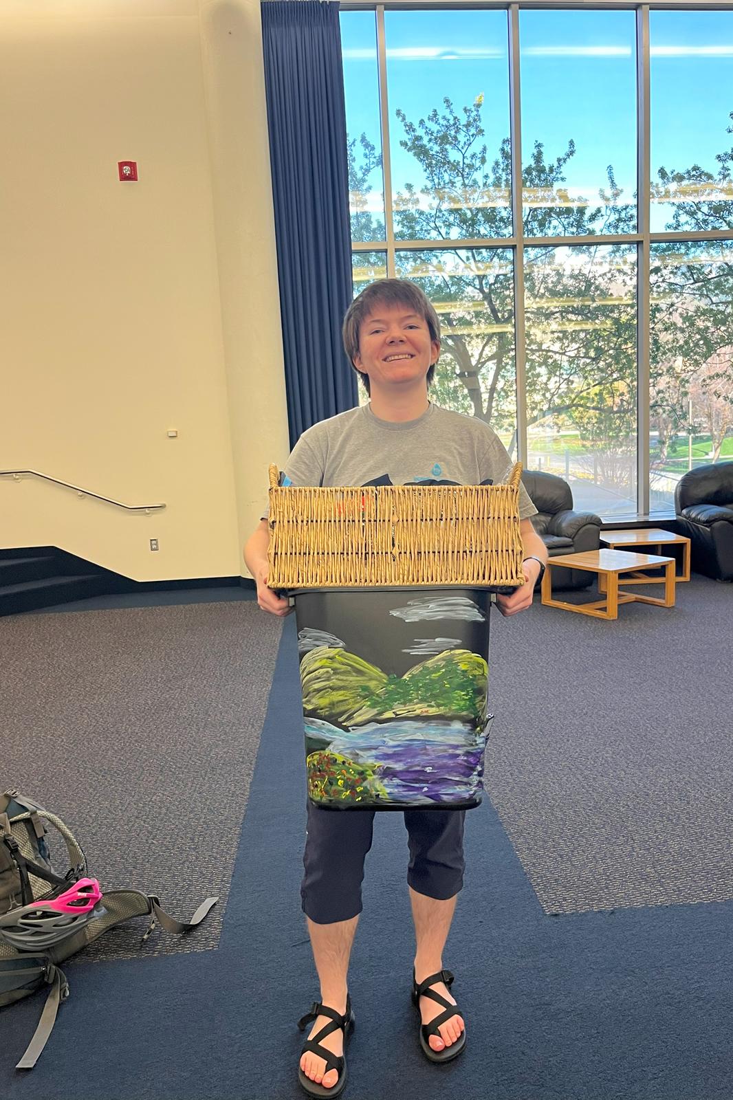 Student holding up a basket and completed recycling bin with a painting of a landscape.