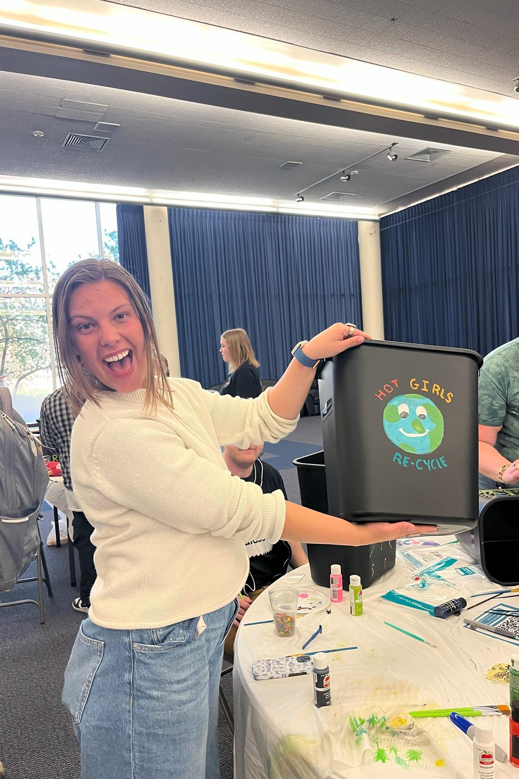 Student holding up their recycling bin with a painting of a smiling Earth surrounded by the words "Hot girls re-cycle".