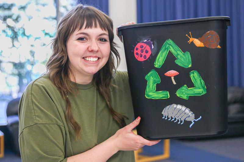 Student holding up their completed recycling bin painted with the recycling symbol, a ladybug, a snail, a mushroom, and a pillbug.