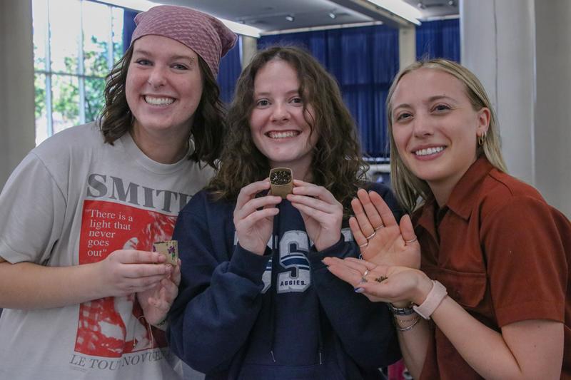 Three students smiling and holding up seeds and two toilet paper tube pots filled with soil.