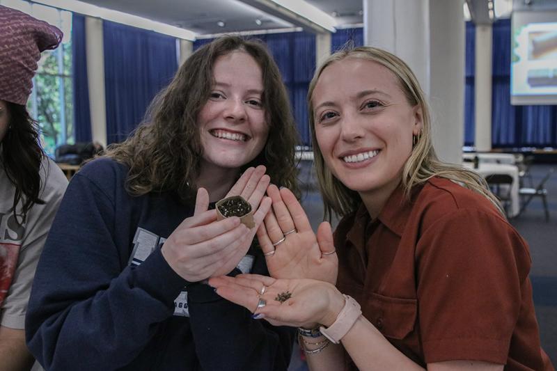 Two students smiling and holding up seeds and a toilet paper tube pot filled with soil.