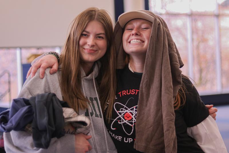 Two students; one holding clothes and smiling, the other wearing clothes on their head and smiling.