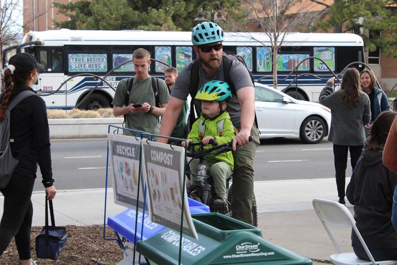 A man and child wearing helmets while riding their bike; compost and recycling bin in front of the bike.