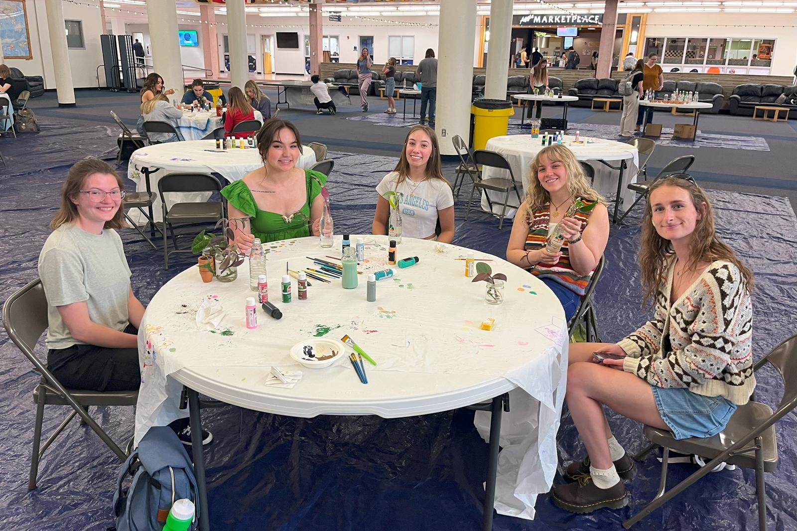 Five students smiling while sitting at a table, planting and painting their reused glass bottles.