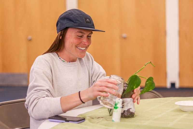 Student sitting at a table and smiling while they water their newly-planted pothos plant in a reused glass jar.