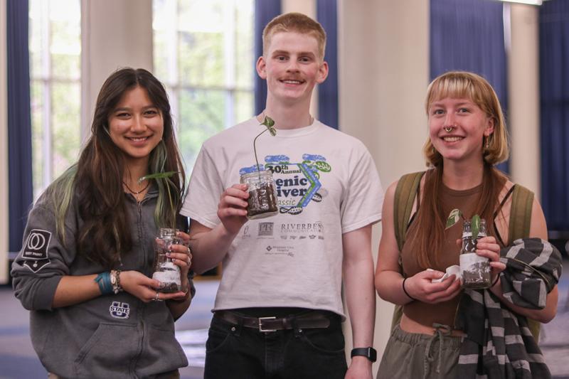 Three students smiling and holding their new plants potted in reused glass jars.