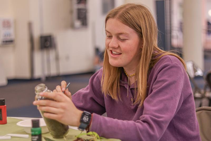 Student sitting at a table and painting a reused glass bottle filled with soil for planting.