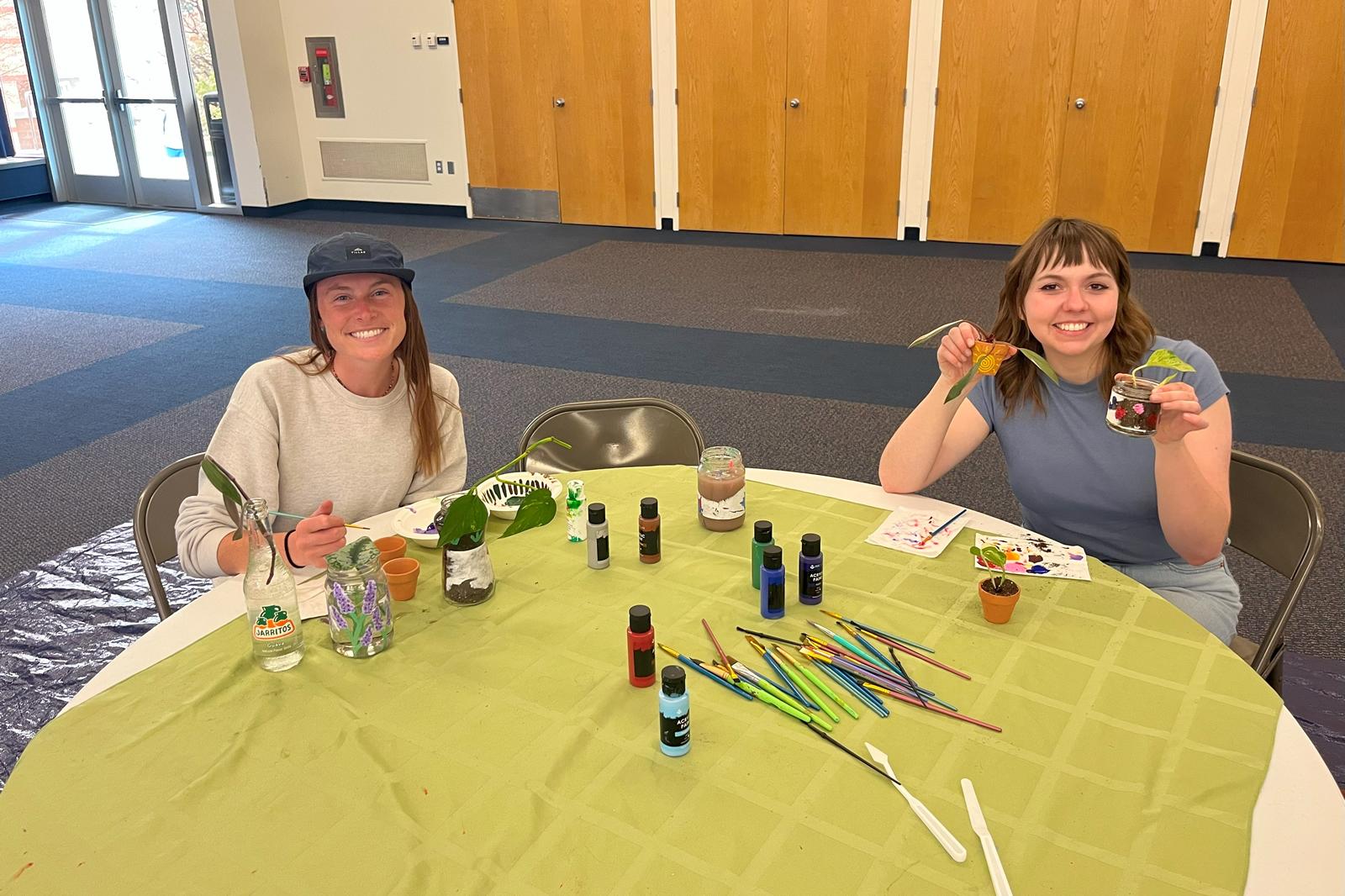Two students smiling while sitting at a table of plants and jars; one student painting the glass jars while the other holds up a small pot and jar they've panted with a sun and berries.