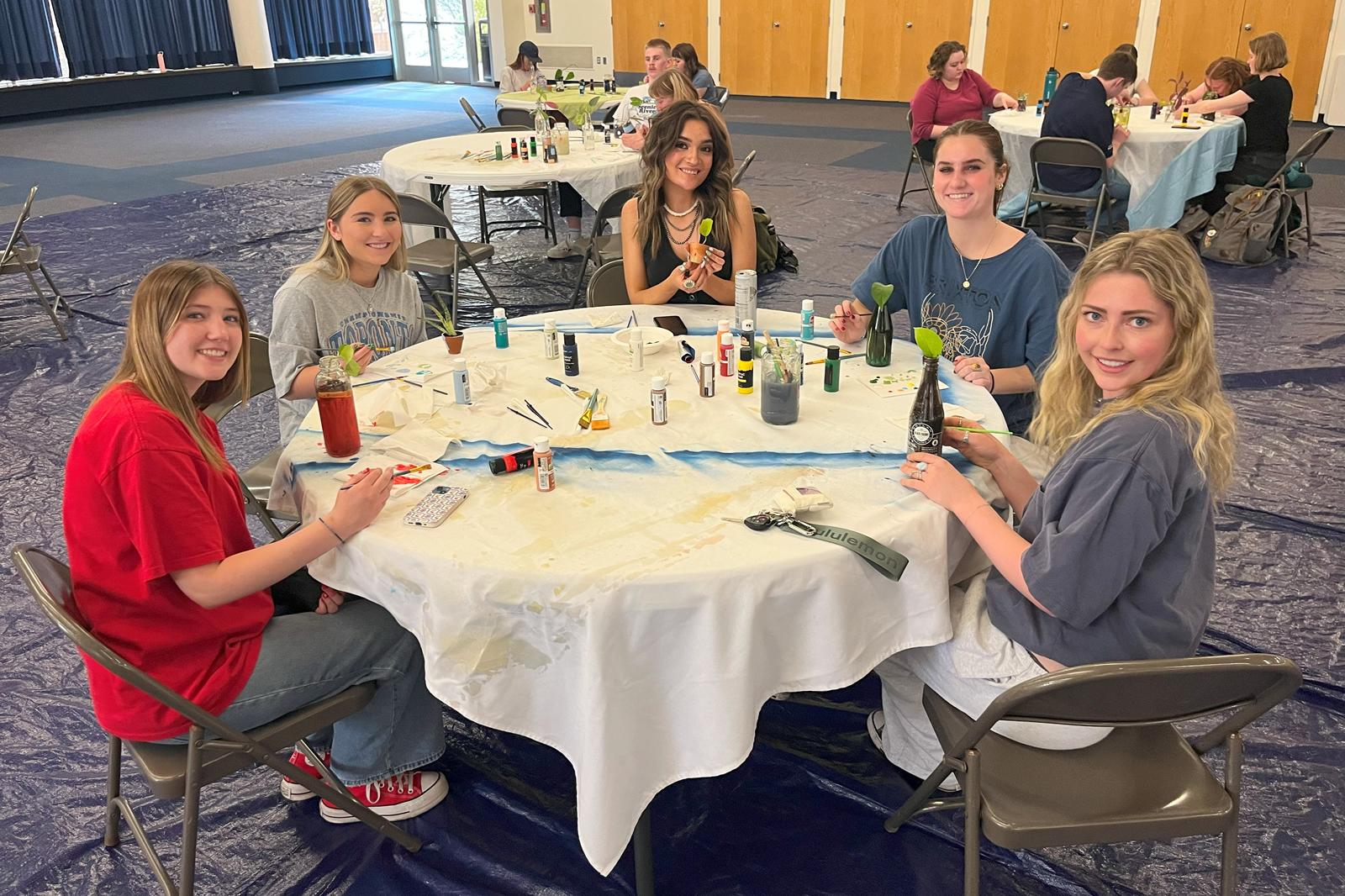 Five students smiling while sitting at a table, painting their reused bottles and small clay pots.