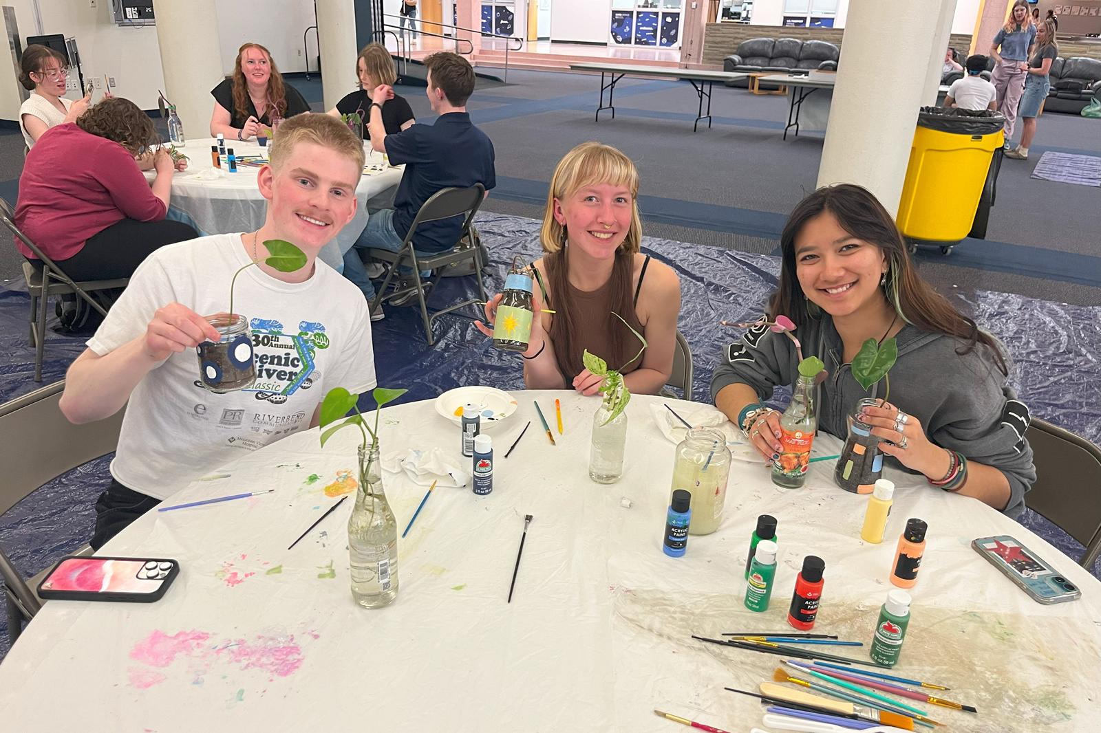 Three students sitting at a table while smiling and holding up their plants in painted glass jars and soil.