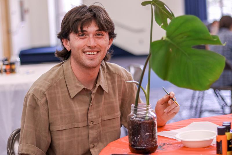 Student sitting at a table and smiling while they paint the reused glass jar they've planted their plant into.