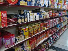 A supermarket aisle with shelves stocked with various packaged food products, including boxes of biscuits, snacks, and tins. A red sale sign on the left side reads “50%,” indicating a discount offer.