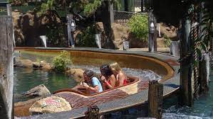 Three people seated in a log-shaped boat on a water ride at an amusement park, surrounded by trees and wooden structures as the ride moves along a winding water channel.