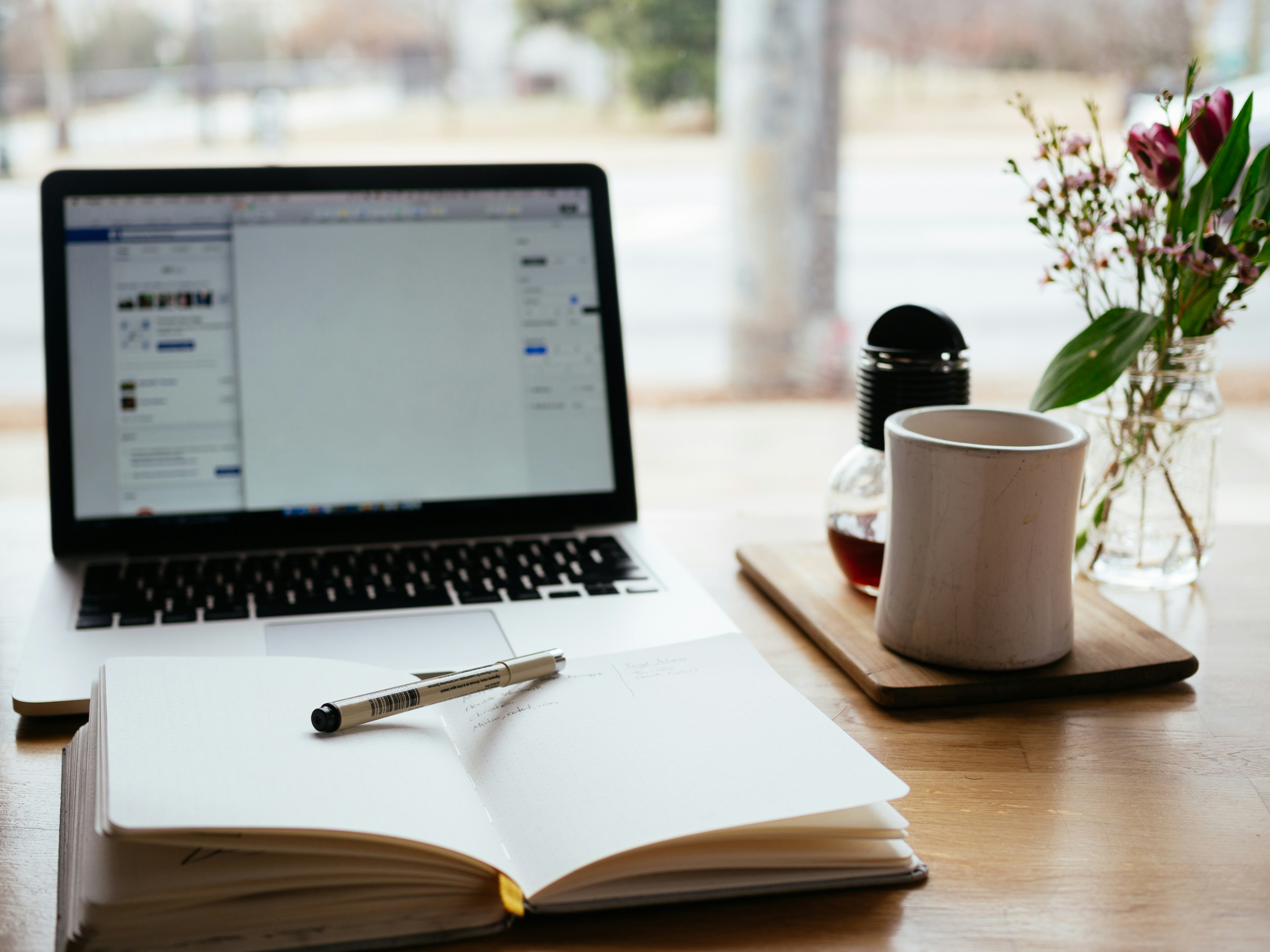 A notebook and laptop on a table next to a beverage and flowers.