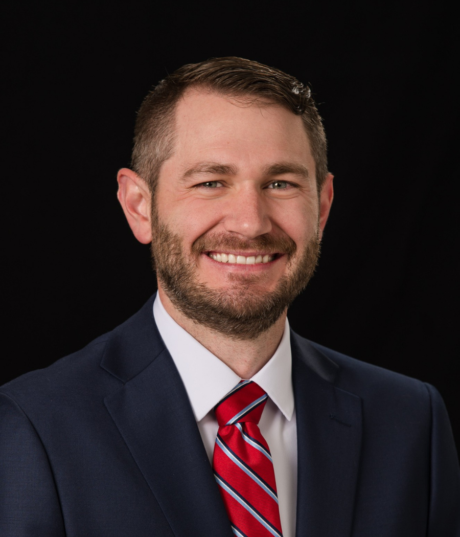 Dane Murray, a bearded man in a dark suit with a red tie, smiling at the camera.