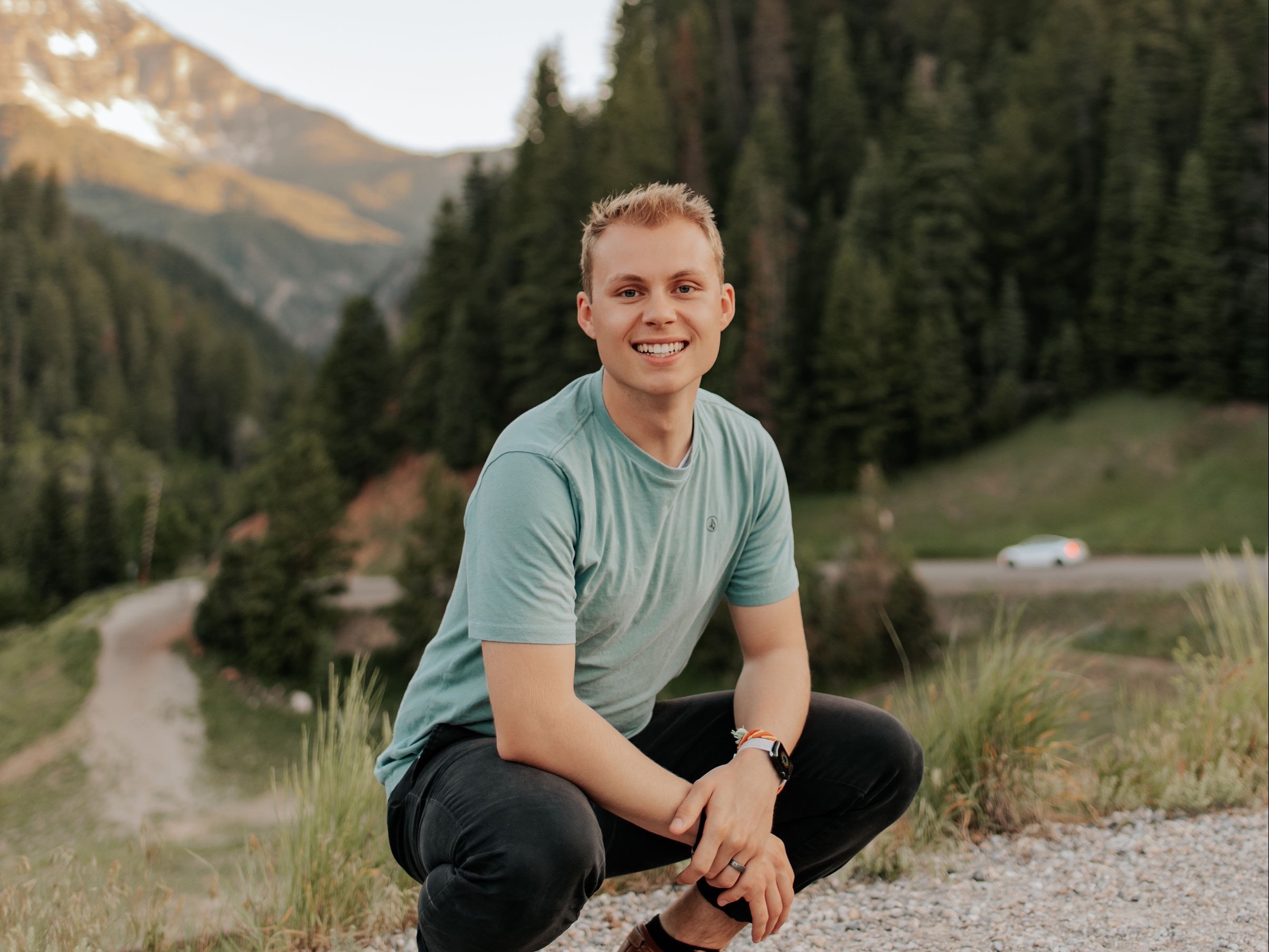 Image of Oliver Heady, a white blonde man, in front of mountains and evergreen trees. Hi smiles at the camera in a squat. He is wearing a turquoise short sleeve shirt, balk pants, and brown leather tennis shoes. 