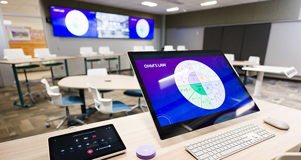 A classroom computer with TVs in the background showing the computer screen.