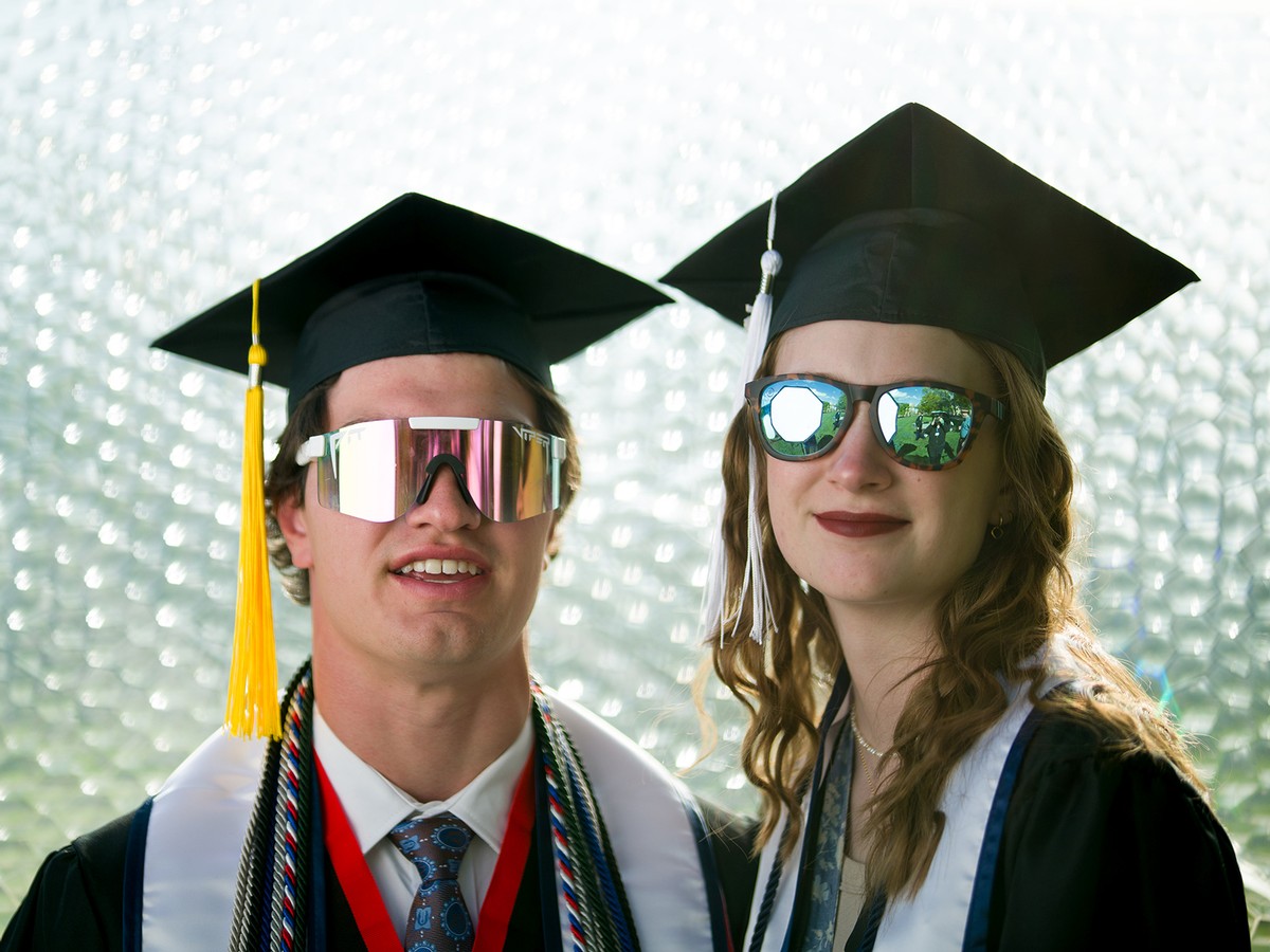 Two graduates in regalia and sunglasses pose for a photo.