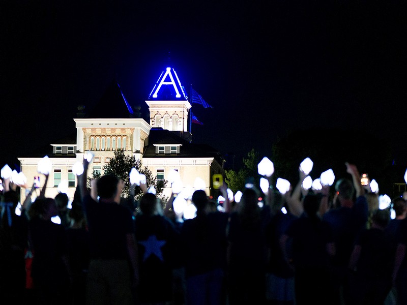 A group of students hold up a Block A light at night, with Old Main in the background
