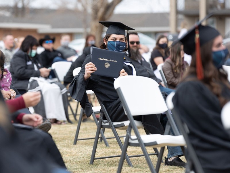 A male USU graduate holds up his diploma while sitting in a chair on the grass.