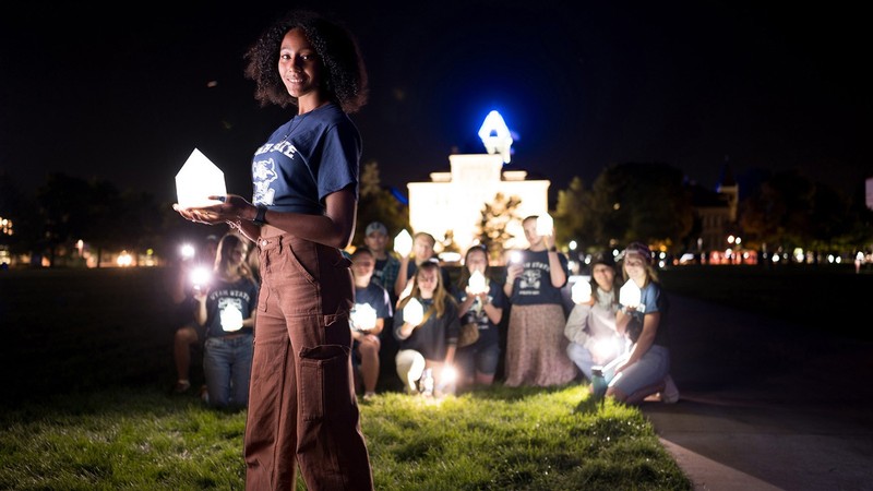 A student holding a lantern shaped like Old Main's tower at night on the USU Logan campus Quad.