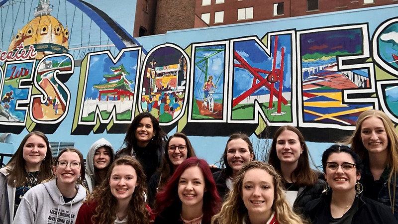College students pose for a photo in front of a mural celebrating Des Moines.