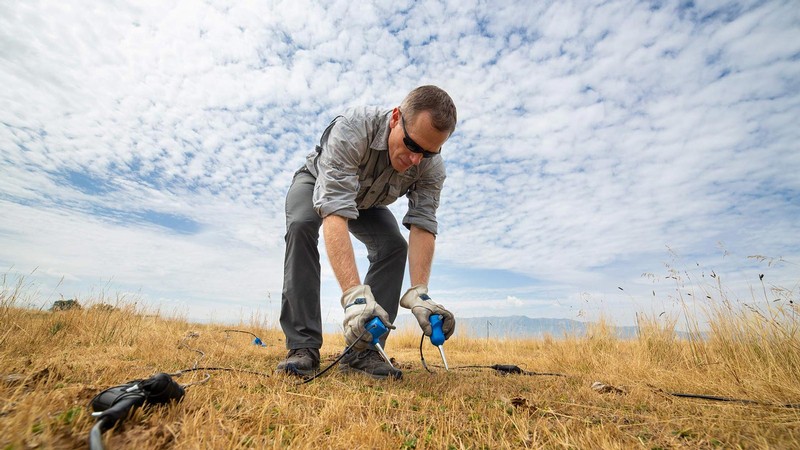 A scientist holding an instrument to the ground in a field.
