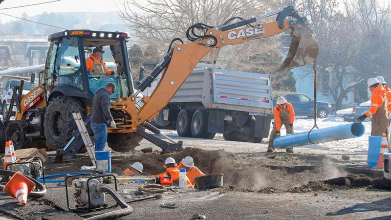 Workers using a backhoe to lift a water pipe into place above a hole in the road.