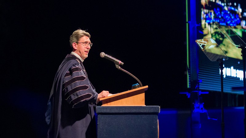 Al Smith wearing graduation robes and speaking at a podium.