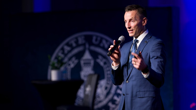 Brad Mortensen holds a microphone and addresses a group of staff at a town hall meeting.