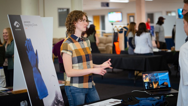 A student at a table explaining paragliding gloves made for a senior exhibit.
