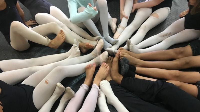 Kids in dance outfits sitting in a circle and putting their feet together.