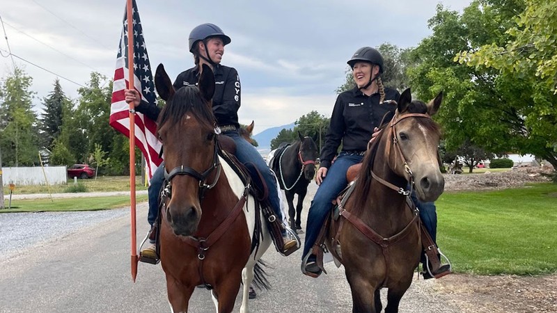 Two women riding horses while one carries an American flag.