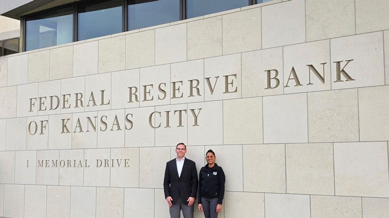 Two people standing by a building. Sign reads: Federal Reserve Bank of Kansas City, 1 Memorial Drive